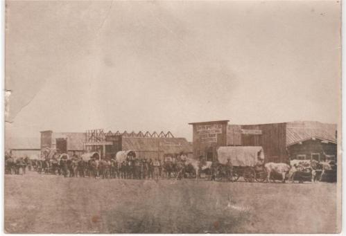 A historic sepia-toned photo of a line of covered wagons and people in front of wooden buildings in a dusty town.