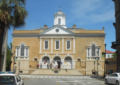 Historic building with a dome, large windows, and flags, set against a clear blue sky. Street view with traffic lights.