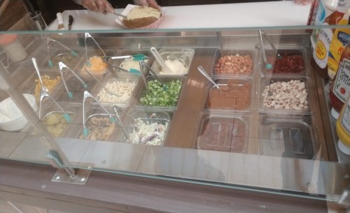 A variety of toppings in containers, including meats, cheeses, and vegetables, displayed behind a glass counter.