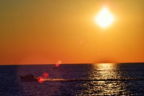 A boat glides across a shimmering ocean at sunset, with a bright sun casting warm hues over the water.