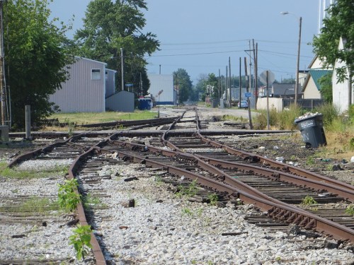 Abandoned railway tracks diverging in an overgrown area, with buildings and a trash bin in the background.