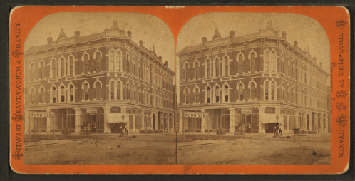 Historic stereoscopic image of a large brick building with storefronts, featuring horse-drawn carriages outside.
