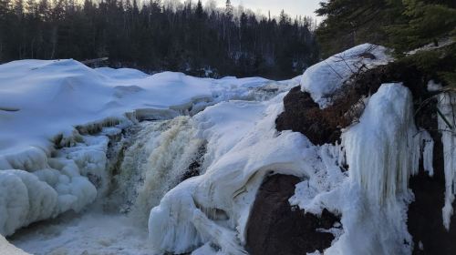 A frozen waterfall surrounded by snow-covered rocks and trees, with icy formations along the edges.
