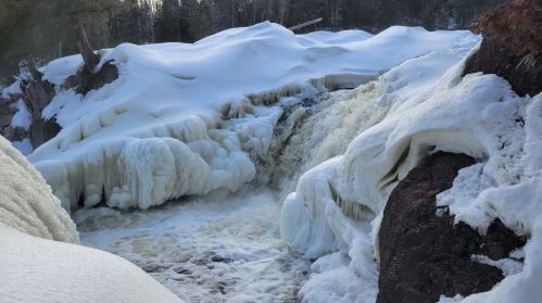 A frozen waterfall surrounded by snow-covered rocks and trees, with rushing water visible beneath the ice.
