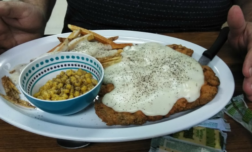 A plate with fried chicken smothered in white gravy, served with fries and a bowl of corn on the side.