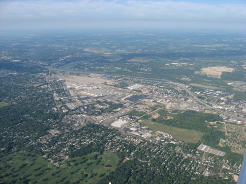 Aerial view of a sprawling urban area with industrial sites, green spaces, and a river in the distance under a blue sky.