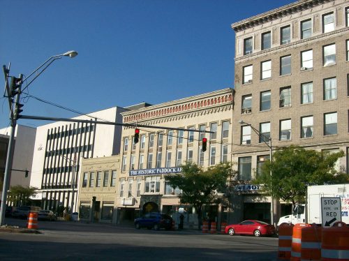 Historic buildings line a city street, with traffic lights and construction barriers in the foreground under a clear blue sky.