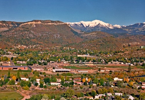 A scenic view of a valley with a small town, surrounded by mountains and snow-capped peaks in the background.