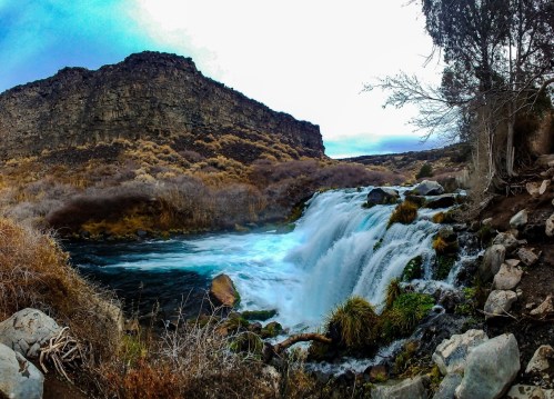 A serene waterfall cascades over rocks, surrounded by rugged cliffs and sparse vegetation under a cloudy sky.