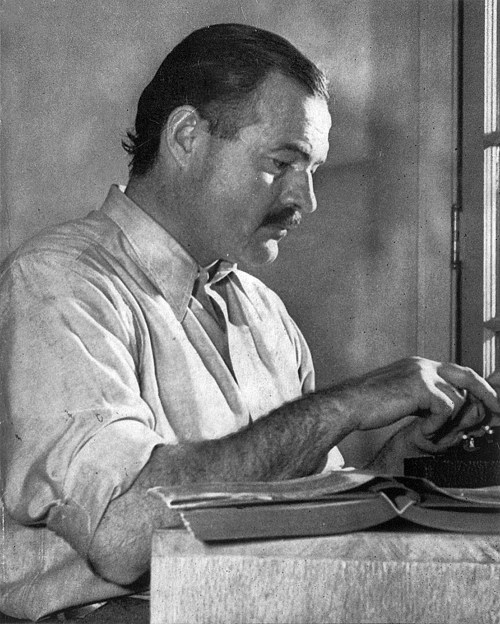 A black and white photo of a man with a mustache, focused on typing at a typewriter in a well-lit room.