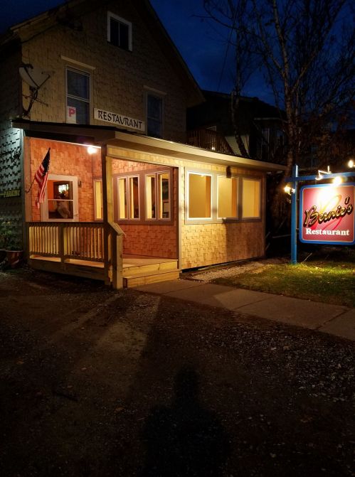 A cozy restaurant exterior at night, featuring wooden siding, a porch, and a colorful sign.