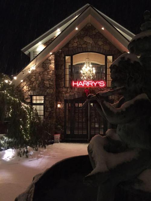 A snowy night scene of a stone building with a neon sign reading "HARRY'S" and a statue playing a flute in the foreground.