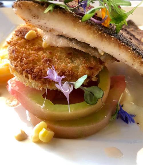 A beautifully plated dish featuring fish, a crispy cake, layered vegetables, and edible flowers.