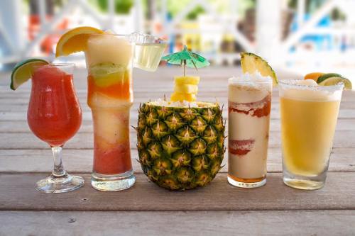 A colorful array of five tropical cocktails, including a pineapple drink, garnished with fruit and umbrellas, on a wooden table.