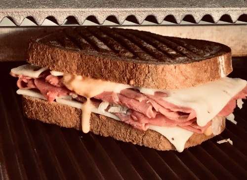 A grilled sandwich with layers of roast beef and melted cheese, served on toasted brown bread.