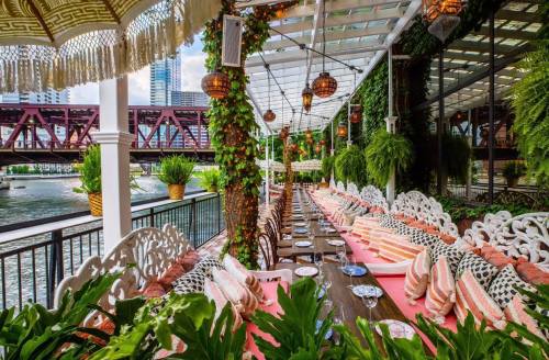A beautifully decorated riverside restaurant with lush greenery, elegant seating, and a view of a bridge.