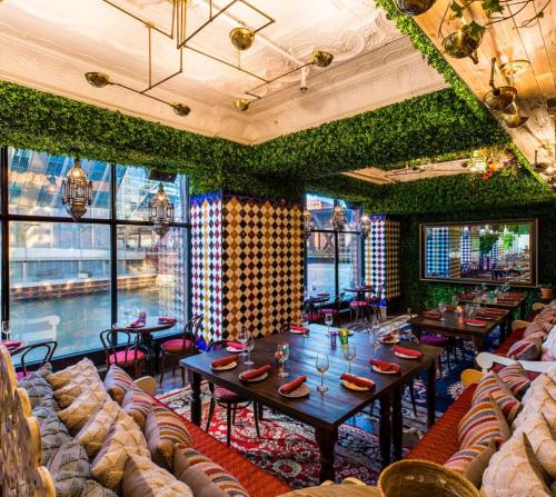 A vibrant restaurant interior with greenery, patterned walls, and a view of the water, featuring wooden tables and colorful decor.