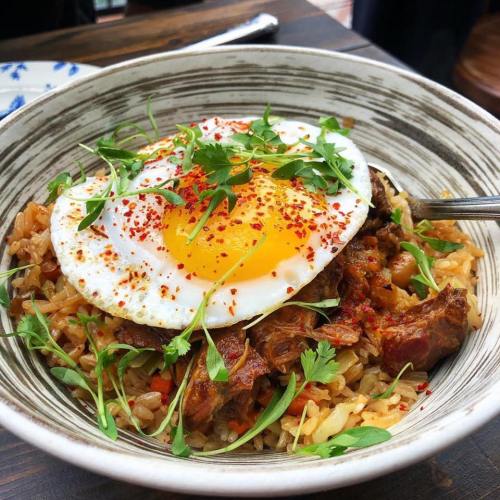 A bowl of rice topped with a fried egg, herbs, and spices, garnished with fresh greens and red pepper flakes.