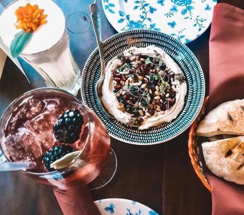 A colorful table setting featuring a bowl of hummus topped with seeds, drinks, and bread on decorative plates.