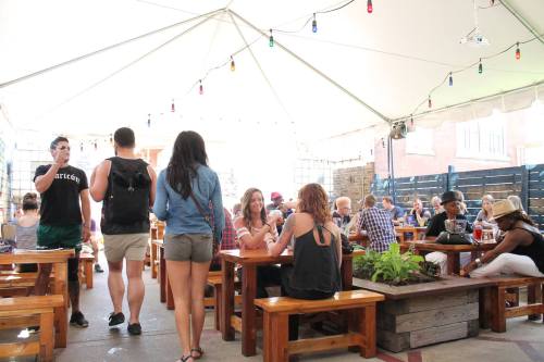 A lively outdoor gathering under a tent, with people socializing at wooden tables and colorful lights overhead.