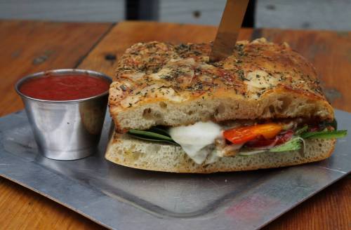 A hearty sandwich with melted cheese, vegetables, and herbs, served with a small cup of marinara sauce on a metal tray.