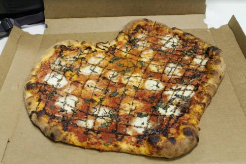 A heart-shaped pizza with a crispy crust, topped with tomato sauce, mozzarella, and fresh basil, in a pizza box.