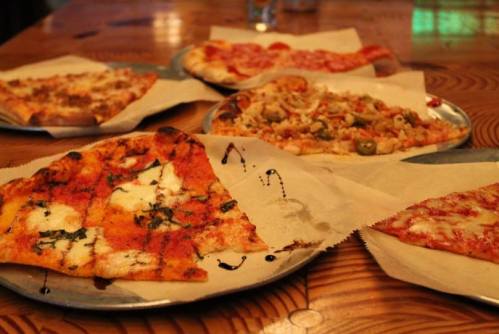 A variety of pizza slices on metal plates, including toppings like cheese, vegetables, and sauce, on a wooden table.