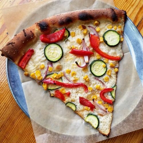 A slice of pizza topped with zucchini, red peppers, corn, and onions on a rustic wooden table.