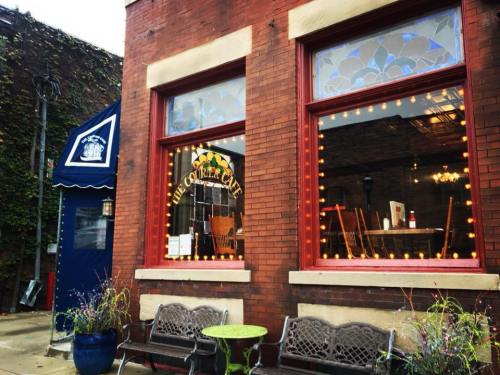 Exterior of The Corner Cafe, featuring large windows, decorative lights, and a blue awning, with benches outside.