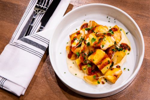A bowl of golden, crispy gnocchi topped with tomato sauce and herbs, served with a fork and knife on a napkin.