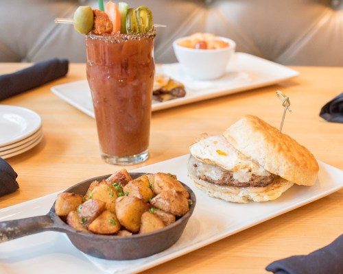 A hearty breakfast featuring a biscuit sandwich with sausage and egg, crispy potatoes, and a colorful cocktail.