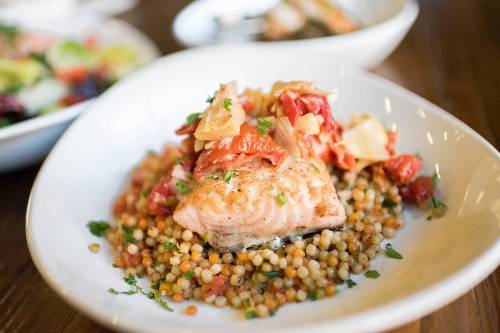A plate of salmon topped with sun-dried tomatoes and artichokes, served over a bed of couscous.
