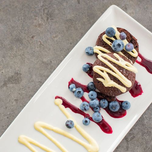 A plate with two chocolate desserts drizzled with white sauce, topped with blueberries and berry sauce.