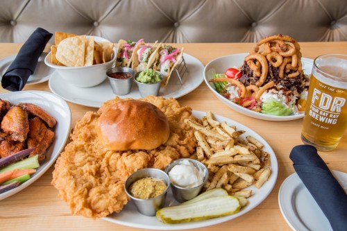 A table spread featuring fried chicken sandwich, fries, wings, tacos, and a craft beer in a casual dining setting.