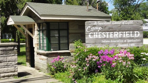 A small stone building with a sign reading "Camp Chesterfield" surrounded by colorful flowers and greenery.