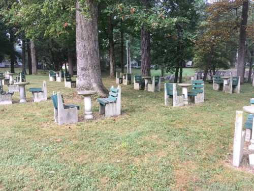 A park scene featuring several stone tables and benches arranged among trees on grassy ground.