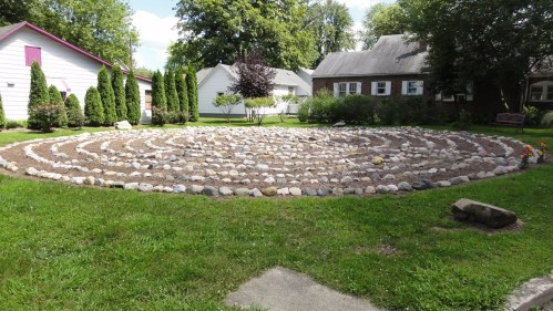 A circular stone labyrinth in a grassy yard, surrounded by trees and houses, under a clear blue sky.