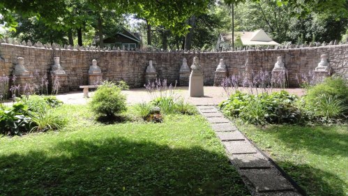 A serene garden with stone sculptures, surrounded by a low stone wall and lush greenery, featuring a pathway through the grass.