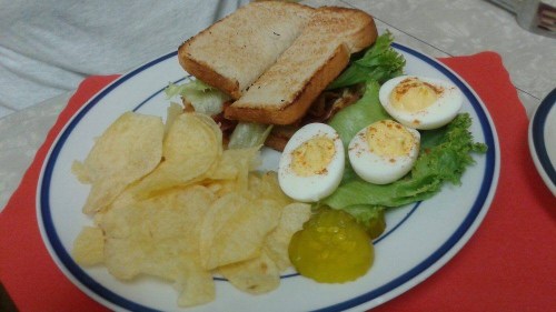 A plate with a sandwich on toasted bread, lettuce, bacon, and two sliced hard-boiled eggs, served with potato chips and pickles.