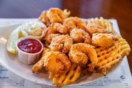 A plate of crispy fried shrimp served with waffle fries, coleslaw, lemon, and a side of dipping sauce.