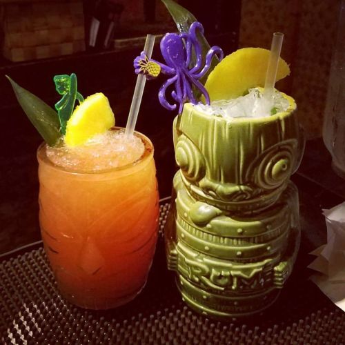 Two colorful cocktails on a bar: one in a tiki glass with an octopus and lime, the other in a tropical glass with a garnish.