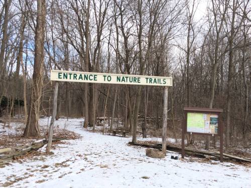 Sign reading "Entrance to Nature Trails" at a snowy forest path with trees in the background.