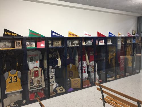 Display of sports memorabilia, including jerseys, trophies, and banners from various schools in glass cases.