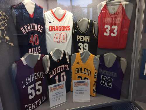 Display of basketball jerseys from various schools, including Heritage Hills, Dragons, Penn, and more, in a showcase.
