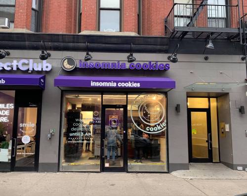 Exterior of an Insomnia Cookies store with a purple awning, featuring a clock logo and large glass windows.