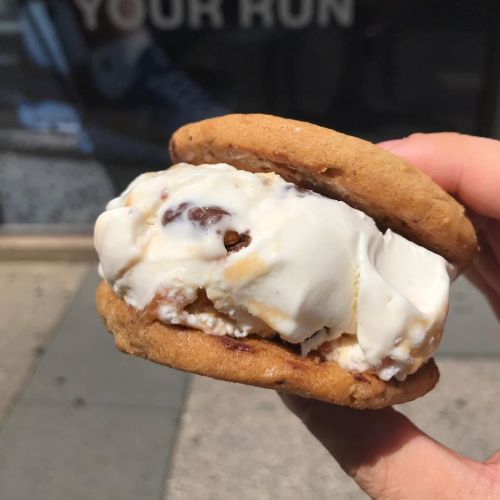 A hand holding a cookie sandwich filled with creamy vanilla ice cream and chocolate chunks, set against a blurred background.