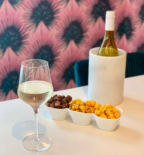 A glass of white wine next to a bottle in a cooler, with bowls of chocolate and cheese popcorn on a table.