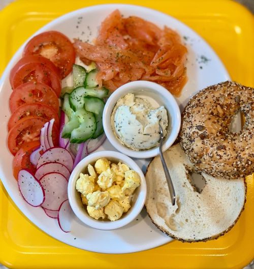 A plate with smoked salmon, sliced tomatoes, cucumbers, radishes, a bagel, and cream cheese, served with popcorn.