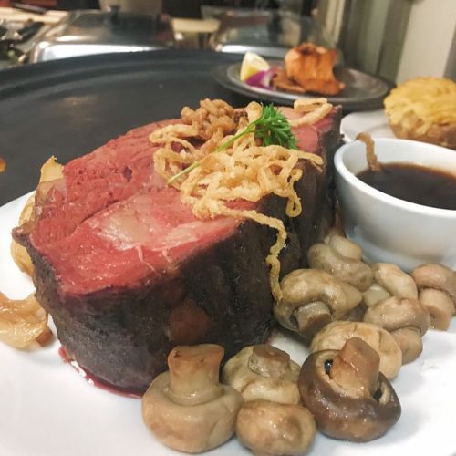 A plate of juicy prime rib with crispy onions, mushrooms, and a side of sauce, garnished with a sprig of parsley.