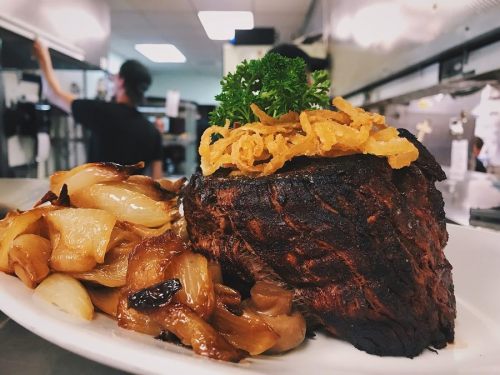 A beautifully plated steak topped with crispy onions, served with caramelized onions in a restaurant kitchen setting.